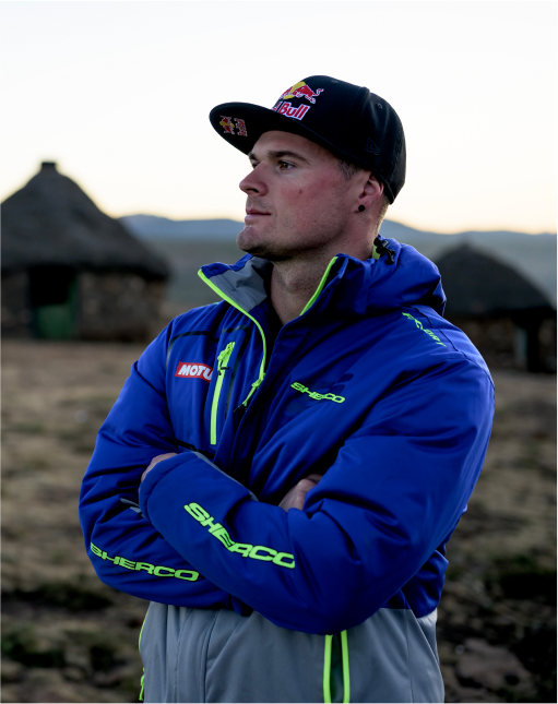 An image of Wade young, in a jacket and cap, looking into the distance to the left of the screen. Behind him is an out of focus dry terrain and there is a hut behind him in the distance.